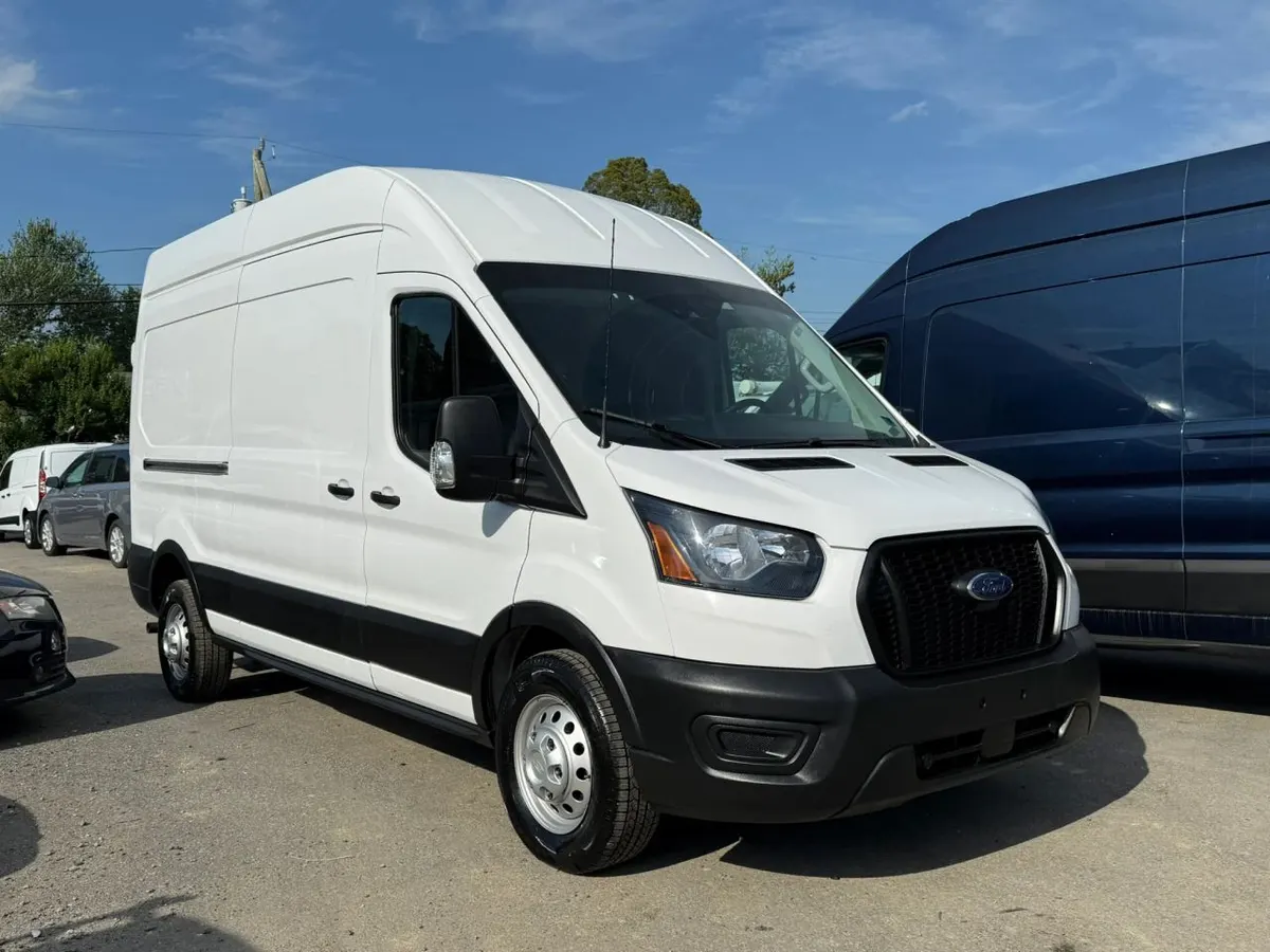 A white Ford Transit high-roof cargo van parked on a gravel lot beside a blue Transit and other vans