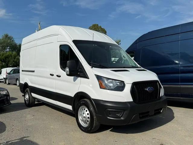 A white Ford Transit high-roof cargo van parked on a gravel lot beside a blue Transit and other vans
